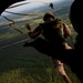 A military paratrooper in camouflage uniform and helmet is exiting an aircraft during a static-line jump exercise, with a yellow static line visible connecting to the aircraft's anchor line cable. A second person in silhouette is visible in the foreground, likely a jumpmaster observing the exit. The terrain below shows dense forested areas with what appears to be an airfield or runway visible in the background.