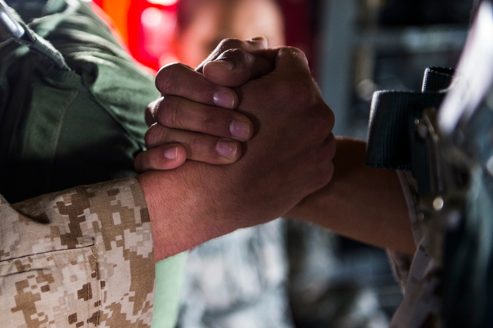 A close-up photograph showing two individuals clasping hands in a firm, interlocked grip. One person is wearing a US Marine Corps MARPAT desert camouflage uniform sleeve, while the other wears a dark tactical wristband or watch strap. The image appears to depict a moment of solidarity, greeting, or camaraderie between military personnel.