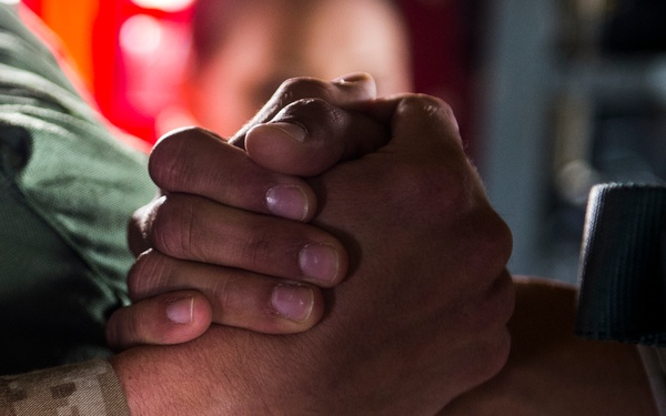 A close-up photograph showing two individuals clasping hands in a firm, interlocked grip. One person is wearing a US Marine Corps MARPAT desert camouflage uniform sleeve, while the other wears a dark tactical wristband or watch strap. The image appears to depict a moment of solidarity, greeting, or camaraderie between military personnel.