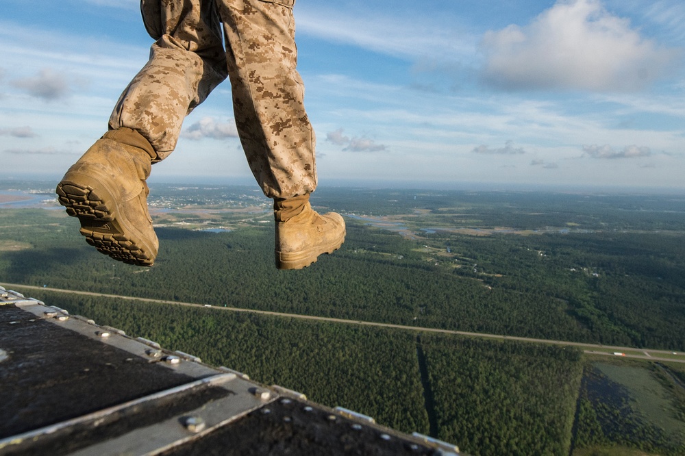 A military personnel wearing digital camouflage (MARPAT pattern) uniform and tan combat boots is stepping off or standing at the edge of an aircraft ramp at significant altitude. The perspective is a low-angle shot from the aircraft's open rear ramp, revealing a vast aerial view of forested terrain with waterways and scattered development below. The image captures the moment just before or during a military parachute jump or airborne operation.