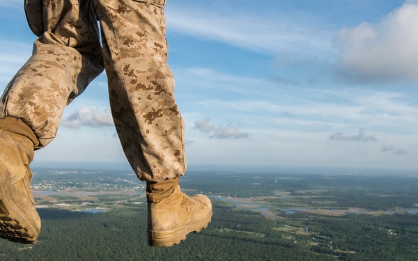 A military personnel wearing digital camouflage (MARPAT pattern) uniform and tan combat boots is stepping off or standing at the edge of an aircraft ramp at significant altitude. The perspective is a low-angle shot from the aircraft's open rear ramp, revealing a vast aerial view of forested terrain with waterways and scattered development below. The image captures the moment just before or during a military parachute jump or airborne operation.