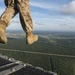 A military personnel wearing digital camouflage (MARPAT pattern) uniform and tan combat boots is stepping off or standing at the edge of an aircraft ramp at significant altitude. The perspective is a low-angle shot from the aircraft's open rear ramp, revealing a vast aerial view of forested terrain with waterways and scattered development below. The image captures the moment just before or during a military parachute jump or airborne operation.