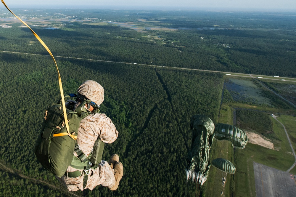 Military paratroopers are jumping from an aircraft over a large forested area with visible airfield infrastructure below. At least two jumpers are visible mid-air, with one in the foreground still connected to a static line (yellow strap) and another further out with a partially deployed green parachute. The terrain below shows dense forest, roads, and what appears to be a military airfield or base.