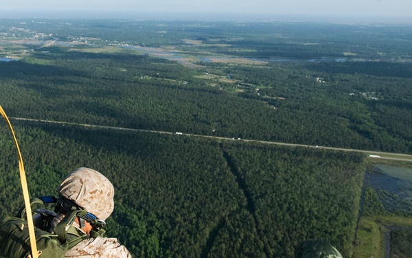 Military paratroopers are jumping from an aircraft over a large forested area with visible airfield infrastructure below. At least two jumpers are visible mid-air, with one in the foreground still connected to a static line (yellow strap) and another further out with a partially deployed green parachute. The terrain below shows dense forest, roads, and what appears to be a military airfield or base.