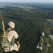 Military paratroopers are jumping from an aircraft over a large forested area with visible airfield infrastructure below. At least two jumpers are visible mid-air, with one in the foreground still connected to a static line (yellow strap) and another further out with a partially deployed green parachute. The terrain below shows dense forest, roads, and what appears to be a military airfield or base.