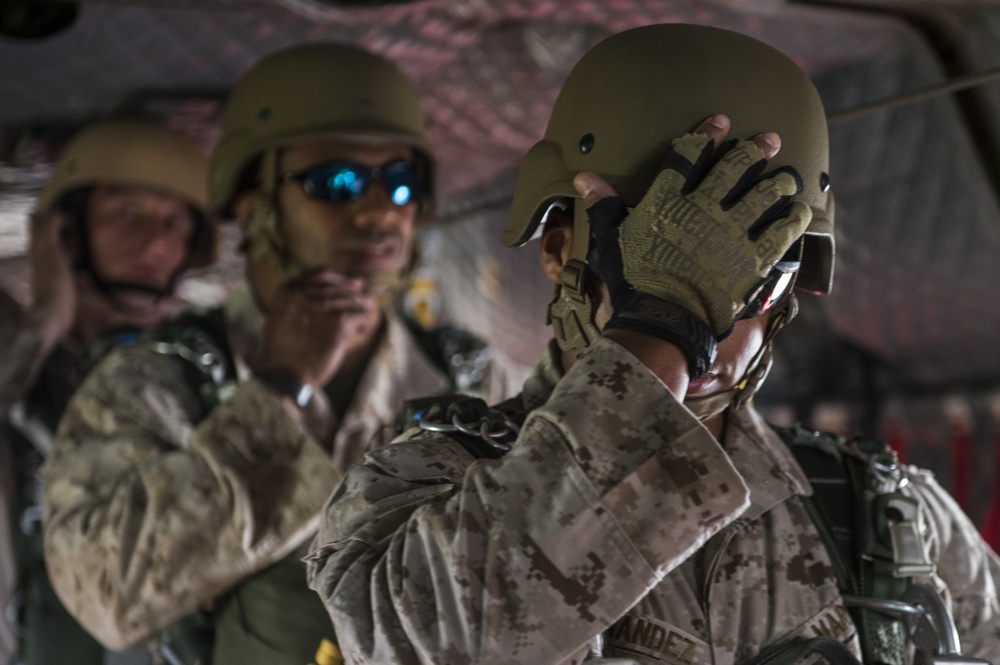 The image shows military personnel, likely U.S. Marines based on the MARPAT (Marine Pattern) camouflage uniforms visible, appearing to be inside a military aircraft or vehicle. The foreground subject is adjusting or securing a tan/olive tactical helmet while wearing fingerless tactical gloves. At least two additional personnel are visible in the background, one wearing blue-tinted sunglasses and tactical gear.