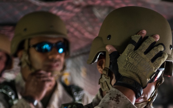 The image shows military personnel, likely U.S. Marines based on the MARPAT (Marine Pattern) camouflage uniforms visible, appearing to be inside a military aircraft or vehicle. The foreground subject is adjusting or securing a tan/olive tactical helmet while wearing fingerless tactical gloves. At least two additional personnel are visible in the background, one wearing blue-tinted sunglasses and tactical gear.