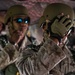 The image shows military personnel, likely U.S. Marines based on the MARPAT (Marine Pattern) camouflage uniforms visible, appearing to be inside a military aircraft or vehicle. The foreground subject is adjusting or securing a tan/olive tactical helmet while wearing fingerless tactical gloves. At least two additional personnel are visible in the background, one wearing blue-tinted sunglasses and tactical gear.