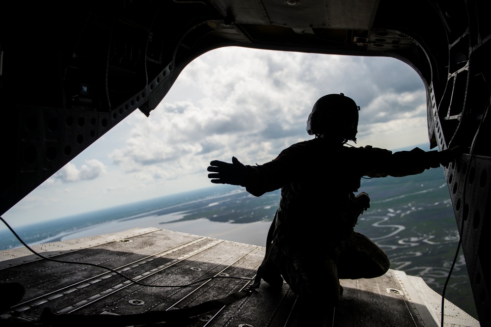 A silhouetted military personnel is crouched at the open rear ramp of a large military helicopter or transport aircraft in flight. The figure is wearing a flight helmet and tactical gear, with one arm extended outward toward the open ramp edge. Below, a landscape featuring rivers, wetlands, and flat terrain is visible under a partly cloudy sky.