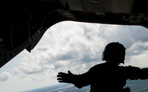 A silhouetted military personnel is crouched at the open rear ramp of a large military helicopter or transport aircraft in flight. The figure is wearing a flight helmet and tactical gear, with one arm extended outward toward the open ramp edge. Below, a landscape featuring rivers, wetlands, and flat terrain is visible under a partly cloudy sky.