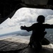 A silhouetted military personnel is crouched at the open rear ramp of a large military helicopter or transport aircraft in flight. The figure is wearing a flight helmet and tactical gear, with one arm extended outward toward the open ramp edge. Below, a landscape featuring rivers, wetlands, and flat terrain is visible under a partly cloudy sky.