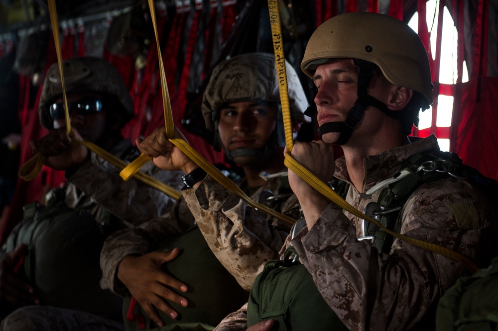 Several military personnel in desert camouflage uniforms are seated inside what appears to be a military transport aircraft. They are holding yellow cargo straps and wearing combat helmets and tactical gear, suggesting preparation for an airborne operation or transport mission. The interior of the aircraft is visible with red netting in the background.