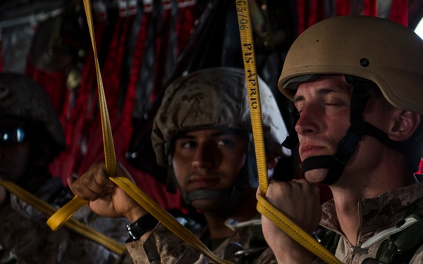 Several military personnel in desert camouflage uniforms are seated inside what appears to be a military transport aircraft. They are holding yellow cargo straps and wearing combat helmets and tactical gear, suggesting preparation for an airborne operation or transport mission. The interior of the aircraft is visible with red netting in the background.