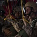 Several military personnel in desert camouflage uniforms are seated inside what appears to be a military transport aircraft. They are holding yellow cargo straps and wearing combat helmets and tactical gear, suggesting preparation for an airborne operation or transport mission. The interior of the aircraft is visible with red netting in the background.