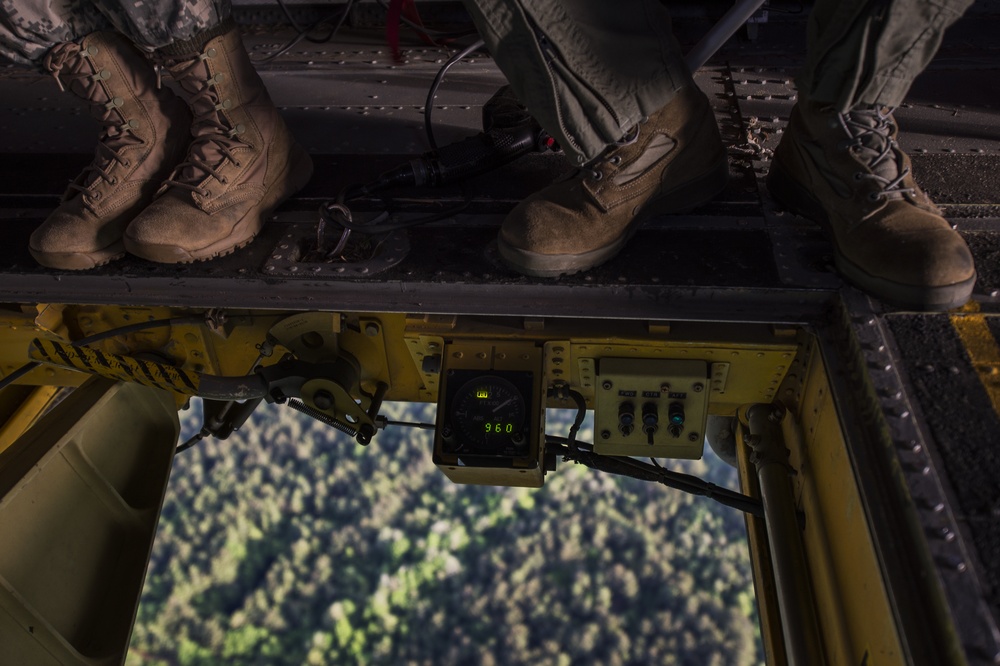 An aerial view taken from inside a military helicopter looking down through an open hatch or ramp. Two personnel in military boots are standing on the edge of the aircraft, with a digital display reading '960' visible on yellow-painted equipment. Dense forest terrain is visible far below through the opening.