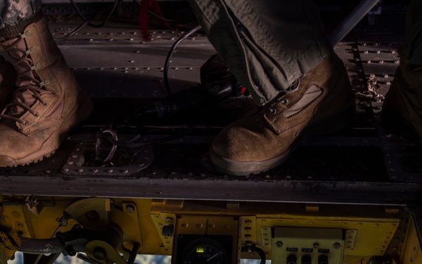 An aerial view taken from inside a military helicopter looking down through an open hatch or ramp. Two personnel in military boots are standing on the edge of the aircraft, with a digital display reading '960' visible on yellow-painted equipment. Dense forest terrain is visible far below through the opening.
