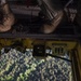 An aerial view taken from inside a military helicopter looking down through an open hatch or ramp. Two personnel in military boots are standing on the edge of the aircraft, with a digital display reading '960' visible on yellow-painted equipment. Dense forest terrain is visible far below through the opening.