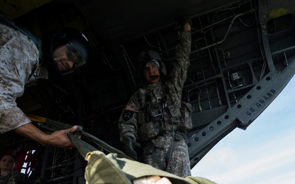 Military personnel are loading or unloading cargo from the rear ramp of a large military transport aircraft, likely a C-130 or similar heavy-lift aircraft. At least three service members in different camouflage patterns (MARPAT and Army ACU) are visible, with one handling a large olive-drab cargo bundle or parachute pack. A 'GO GUARD' placard is visible on the aircraft's interior ramp structure.