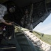 Military personnel are loading or unloading cargo from the rear ramp of a large military transport aircraft, likely a C-130 or similar heavy-lift aircraft. At least three service members in different camouflage patterns (MARPAT and Army ACU) are visible, with one handling a large olive-drab cargo bundle or parachute pack. A 'GO GUARD' placard is visible on the aircraft's interior ramp structure.