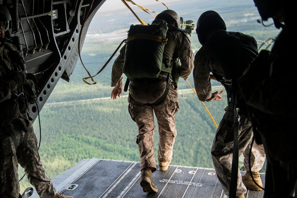 Military personnel are conducting a static-line parachute jump from the open rear ramp of a military cargo aircraft, likely a CH-47 Chinook or C-130. One soldier in camouflage uniform and full parachute pack is positioned at the edge of the ramp, about to jump, while others observe from inside the aircraft. The terrain visible below consists of dense forested areas with some cleared land.