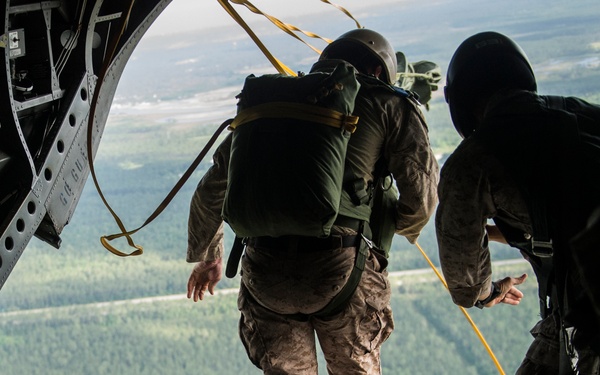 Military personnel are conducting a static-line parachute jump from the open rear ramp of a military cargo aircraft, likely a CH-47 Chinook or C-130. One soldier in camouflage uniform and full parachute pack is positioned at the edge of the ramp, about to jump, while others observe from inside the aircraft. The terrain visible below consists of dense forested areas with some cleared land.