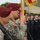 A military ceremony featuring U.S. Army Airborne soldiers in ACU (Army Combat Uniform) wearing maroon berets, standing at attention with M4 carbine rifles. They are accompanied by what appear to be foreign military personnel in dark blue dress uniforms, suggesting a joint or multinational ceremonial event. A guidon or unit flag with gold fringe is being carried by one of the soldiers.