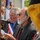 A bearded man wearing sunglasses and a dark suit with a red tie is speaking at a microphone while holding papers, appearing to give a speech at a formal outdoor ceremony. Behind him stands another man wearing a tricolor sash (red, white, blue), suggesting a French official or dignitary. The scene includes ceremonial flags and banners, indicating a commemorative or civic event.