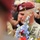 A U.S. Army officer wearing a maroon beret and ACU (Army Combat Uniform) is holding a bouquet of colorful flowers, with his head bowed in what appears to be a solemn or emotional moment. The maroon beret with a flash insignia identifies him as a paratrooper, likely from the 82nd Airborne Division. He is surrounded by a crowd of people in a ceremonial or commemorative setting.