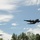 Two military turboprop transport aircraft are flying in formation over a tree-lined area under a partly cloudy blue sky. The foreground aircraft is clearly visible and identifiable as a large four-engine turboprop, consistent with a C-130 Hercules type, while a second aircraft of the same type is visible in the background at greater distance. The image is taken from a low ground-level perspective looking upward.