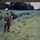 An elderly man stands in a rural, marshy field beside a small waterway or drainage ditch lined with tall reeds and grass. He is wearing an olive drab military-style uniform adorned with multiple patches, medals, and insignia, including a visible American flag patch and a Red Cross armband. The setting appears to be a commemorative or memorial visit, possibly in Normandy or another WWII battlefield site.
