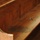The image shows a close-up view of a wooden church pew or bench with visible wear and damage. The seating surface displays multiple dark stains or moisture damage marks, and the wood finish shows significant aging and deterioration. The pew features traditional paneled backrest construction with curved end supports.