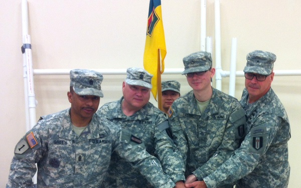 Four U.S. Army soldiers in ACU (Army Combat Uniform) are gathered around a table cutting a sheet cake celebrating the United States Army's 239th birthday. Three soldiers are jointly holding a knife to cut the cake while a fourth stands beside them. A military unit flag (yellow and blue) is visible in the background.