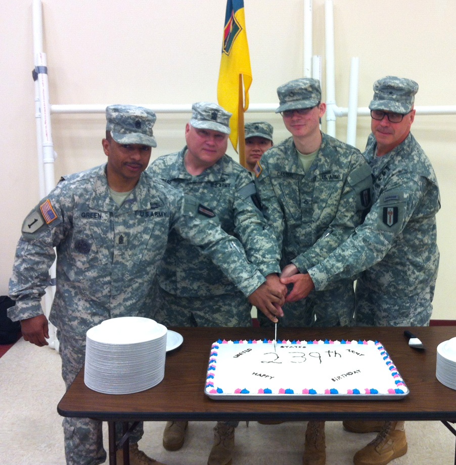 Four U.S. Army soldiers in ACU (Army Combat Uniform) are gathered around a table cutting a sheet cake celebrating the United States Army's 239th birthday. Three soldiers are jointly holding a knife to cut the cake while a fourth stands beside them. A military unit flag (yellow and blue) is visible in the background.