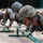 ROK soldiers, US Marines strap in for Mountain Warfare Training Course Multiple soldiers in camouflage uniforms are performing a low-crawl exercise under horizontal bars mounted on green metal stands, likely part of a military obstacle course or physical training drill. At least two distinct camouflage patterns are visible, suggesting a joint or multinational military training exercise. The foreground soldier wears USMC MARPAT digital camouflage and carries a full pack.