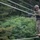 ROK soldiers, US Marines strap in for Mountain Warfare Training Course A military service member in full combat uniform and helmet navigates a high rope obstacle course, balancing on a lower rope while gripping two lateral guide ropes. The individual is wearing MARPAT (Marine pattern) camouflage, suggesting affiliation with the United States Marine Corps. The setting appears to be an outdoor military training facility surrounded by dense green foliage.