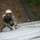 ROK soldiers, US Marines strap in for Mountain Warfare Training Course A U.S. Marine in MARPAT digital camouflage uniform is crouching on what appears to be a sloped metal surface, handling rope or cable rigging. The Marine is wearing a cover (cap), sunglasses, and gloves while working with what appears to be a twisted rope or cable system laid across the surface. The background shows a hillside with vegetation and a dark pipe or post.