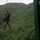 ROK soldiers, US Marines strap in for Mountain Warfare Training Course A military service member in full combat uniform and helmet is suspended from a rope attached to a green steel training tower structure. The individual appears to be conducting a rappelling or rope descent exercise against a backdrop of densely forested green hills. The overcast sky and lush vegetation suggest a temperate, possibly East Asian training environment.