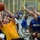 A wheelchair basketball game is in progress at what appears to be the Warrior Games, a military adaptive sports competition. A player in a yellow jersey is attempting a layup or shot while defenders in blue 'Warrior Games' jerseys attempt to block. The event is held in an indoor gymnasium with an 'ARMY' banner visible in the background and spectators watching courtside.