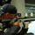 A male shooter is aiming a Feinwerkbau precision air rifle at an indoor shooting range. He is wearing an Under Armour cap and a black and yellow shooting jacket, using proper competitive shooting form. The background shows an indoor range with multiple shooting lanes and target holders.