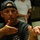 A man wearing a navy blue baseball cap worn backwards and a dark t-shirt is gesturing with both hands, appearing to be explaining or demonstrating something. His cap reads 'HONOR AND' on the front brim, and his shirt displays an 'RSLC' logo patch. A second person in a white t-shirt is visible but blurred in the background.