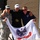 Three men pose together outdoors in front of a brick building, holding a United States Army flag. The man in the center is wearing a medal and a cap with 'Warrior' text, flanked by a soldier in Army Combat Uniform on the left and a man in a navy polo shirt on the right. The image appears to be taken at a military or veteran-related event.