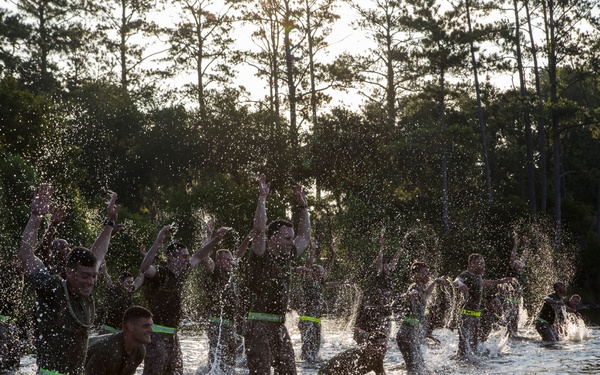 A large group of individuals wearing dark military-style uniforms and distinctive neon green belts/cummerbunds are wading and splashing through a body of water, with arms raised in celebration or exertion. The scene appears to be a military training exercise or graduation event taking place at dusk or dawn, with backlit trees visible in the background. The dynamic water splashing and celebratory body language suggest a milestone completion or team-building activity.