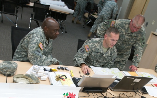 Military personnel in US Army ACU (Army Combat Uniform) are gathered around a table during the Golden Coyote 2014 exercise, specifically at Station 5 for SGLV/DD 93 processing. The scene takes place in an indoor briefing or operations room with multiple soldiers working at laptops and reviewing documents. A placard identifies this as the 265th Support Group (REB) Headquarters station.