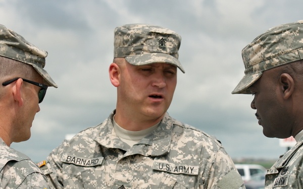 Three U.S. Army soldiers in ACU (Army Combat Uniform) digital camouflage are engaged in a discussion outdoors. The center soldier, identified by name tape as 'BARNARD,' appears to be a senior NCO based on rank insignia visible on his cap, and is holding what appears to be a notepad or small booklet. The soldiers on the left and right are identified by name tapes as 'LOWE' and 'HUNT' (partially visible) respectively.