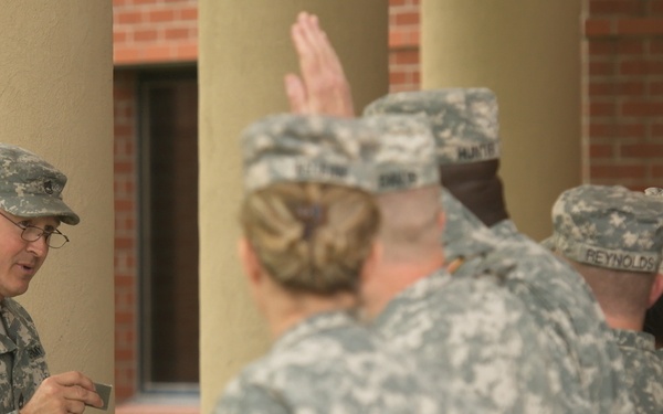 A U.S. Army soldier, likely an officer or NCO, stands facing a group of soldiers who have their right hands raised, suggesting an oath-taking or swearing-in ceremony. The soldier on the left is holding what appear to be small boxes, possibly containing medals, coins, or insignia. The setting appears to be an outdoor covered walkway adjacent to a brick building.