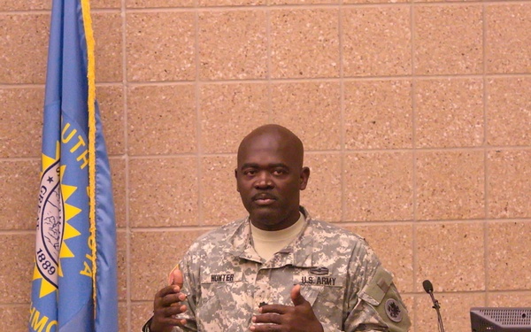 A U.S. Army soldier named Hunter is delivering a briefing or presentation at a podium to an audience of fellow soldiers in ACU (Army Combat Uniform) camouflage. A South Dakota state flag is visible in the background to the left. The setting appears to be a military classroom or briefing room with cinder block walls.