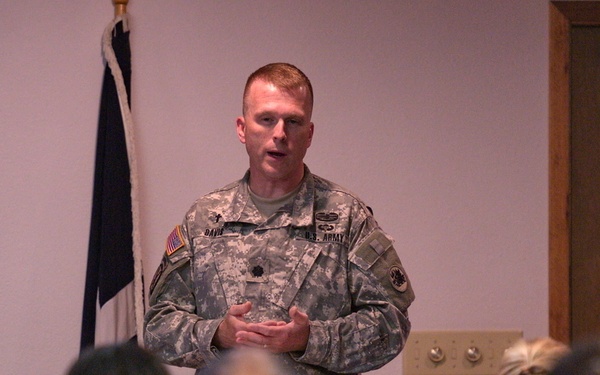 A U.S. Army soldier in ACU (Army Combat Uniform) is addressing a small group of seated military personnel in what appears to be a chapel or briefing room. The speaker's name tape reads 'DAVIS' and he wears a U.S. Army tape with an American flag patch on his left shoulder. The audience, also in military uniforms, is seated in chairs facing the speaker.