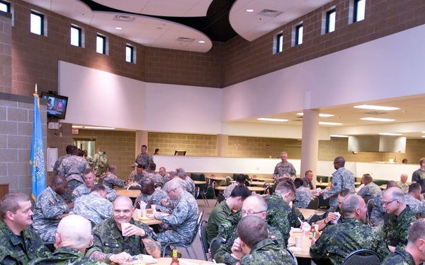 A large group of military personnel from multiple nations are gathered in what appears to be a military dining facility (DFAC/mess hall). Soldiers wearing Canadian CADPAT camouflage uniforms are prominently visible alongside U.S. Army personnel in ACU pattern uniforms, suggesting a multinational military exercise or joint training event. The setting is a modern, well-lit cafeteria-style facility with round and rectangular tables.
