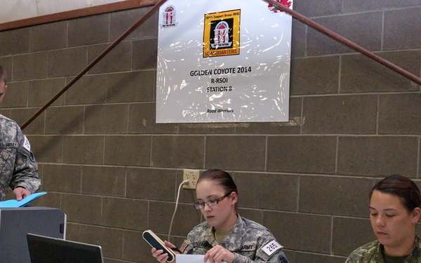 Three military personnel are working at a processing station during the Golden Coyote 2014 R-RSOI exercise, Station 8. Two U.S. Army soldiers and one Canadian Armed Forces soldier are seated and standing at a table equipped with a laptop, printer, and extensive paperwork. The sign on the wall identifies this as 'Golden Coyote 2014 R-RSOI Station 8 - Road Warriors.'