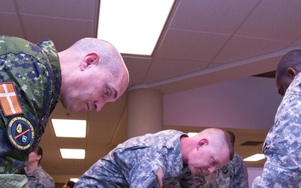 Military personnel from at least two different nations are gathered around a table in an indoor facility, writing or signing documents. The soldier on the left wears a Danish flag patch and what appears to be a Danish Home Guard unit patch, while the other soldiers wear US Army ACU (Army Combat Uniform) pattern camouflage. This appears to be a multinational military exercise or administrative event.